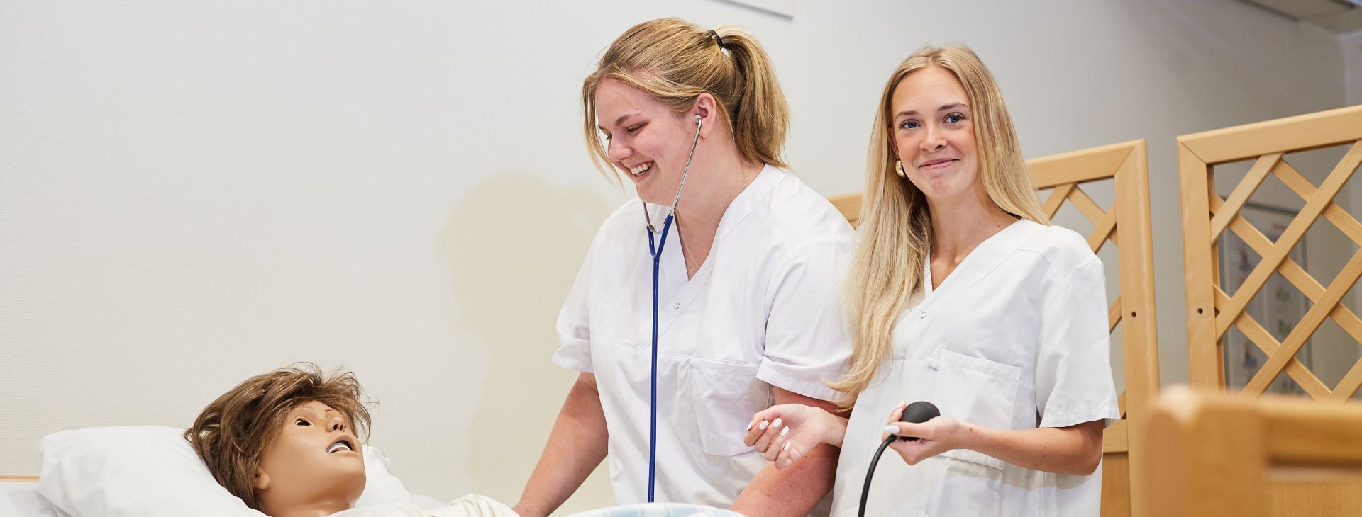 Två tjejer med sjukvårdskläder, som står vid en säng.