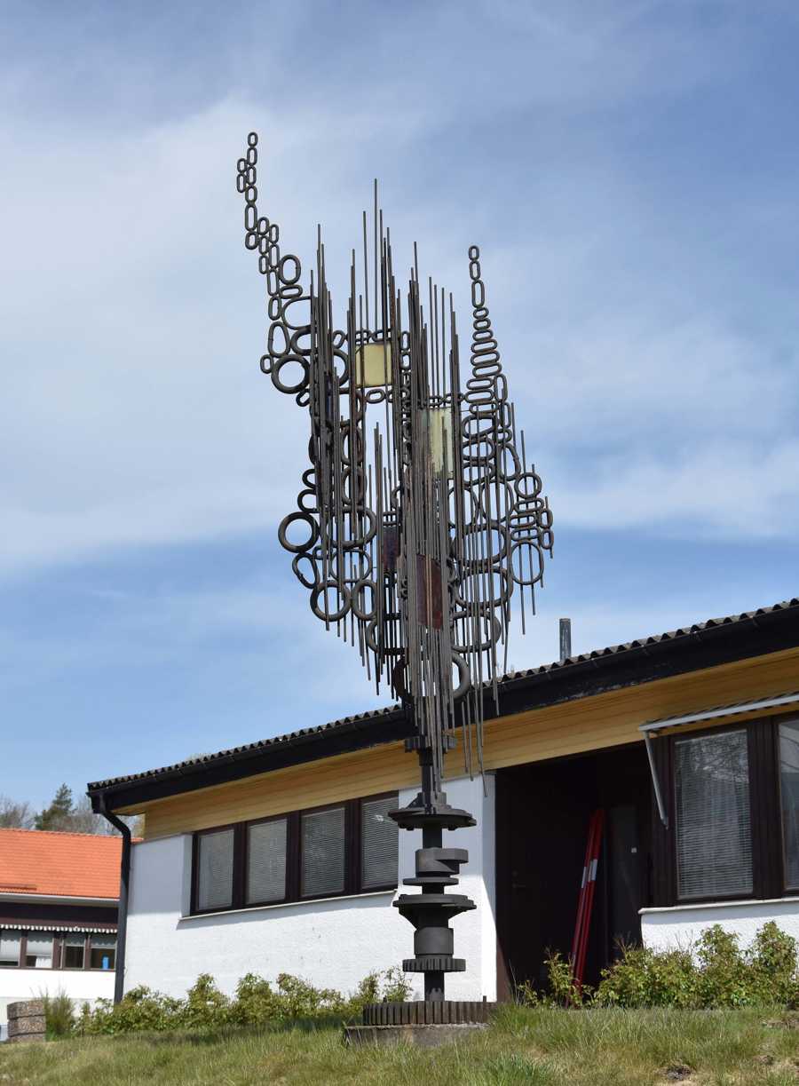 Skulptur gjord av kättingsringar, metallstavar och glas på ett fundament av kugghjul. 