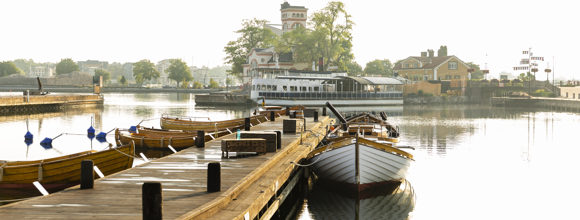 Bild på en träbrygga och en träbåt med utsikt över vatten och byggnader i bakgrunden.