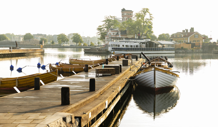 Bild på en träbrygga och en träbåt med utsikt över vatten och byggnader i bakgrunden.