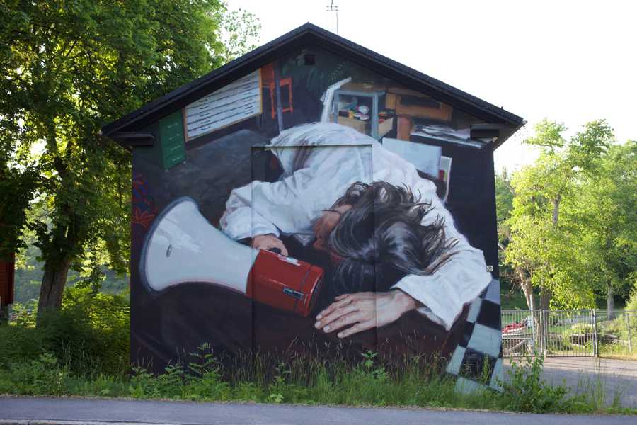 Väggmålning föreställande en person i vit målarrock som ligger över ett bord och håller i en röd och vit megafon.