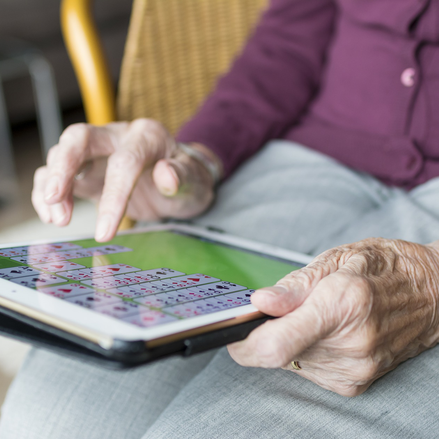 Bilden visar en äldres kvinnas händer. Hon har en surfplatta i sitt knä och spelar kortspel på den.
