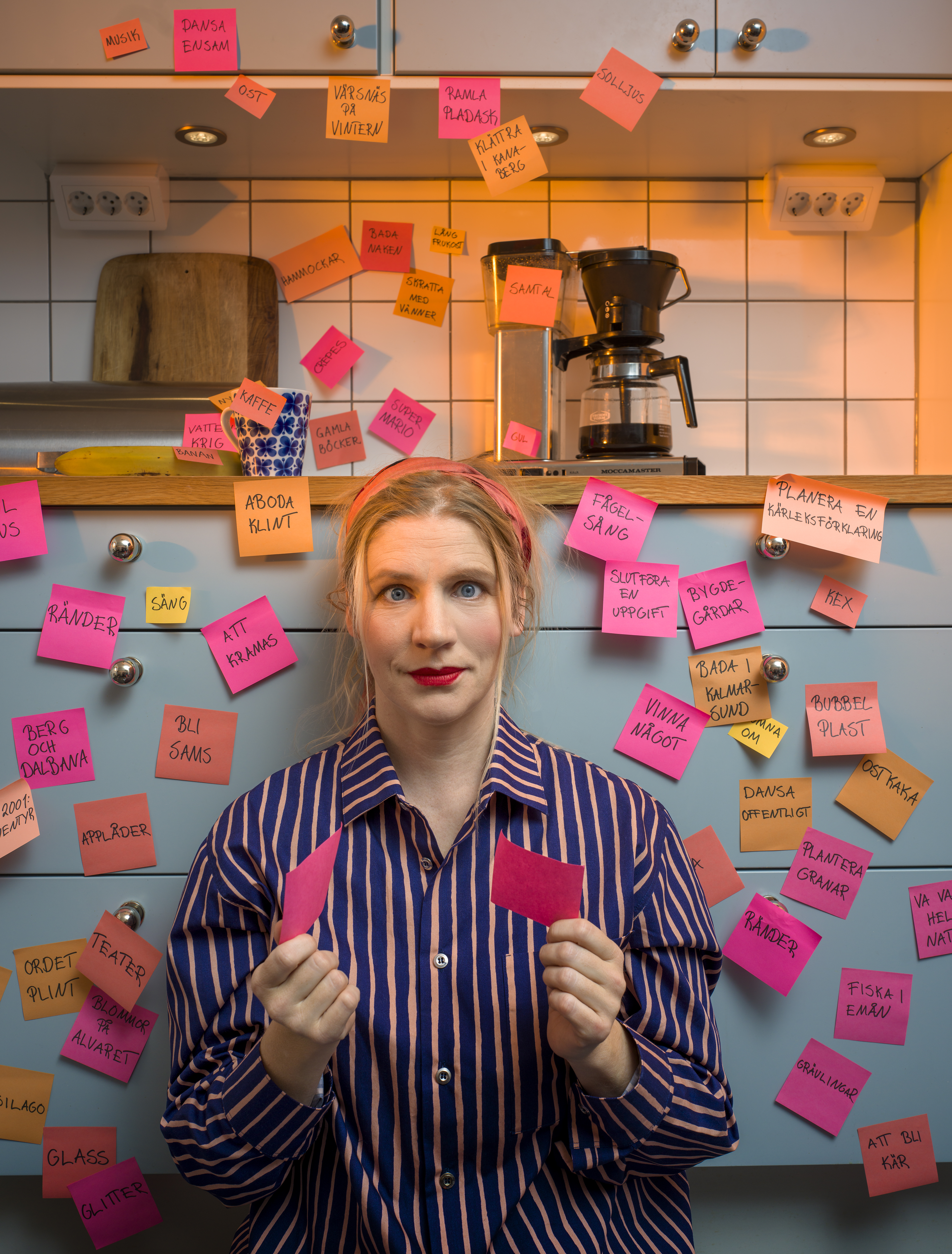 Bildtolkning: En kvinna sitter på golvet i ett kök omgiven av post-it-lappar. Hon har också post-it-lappar i händerna.