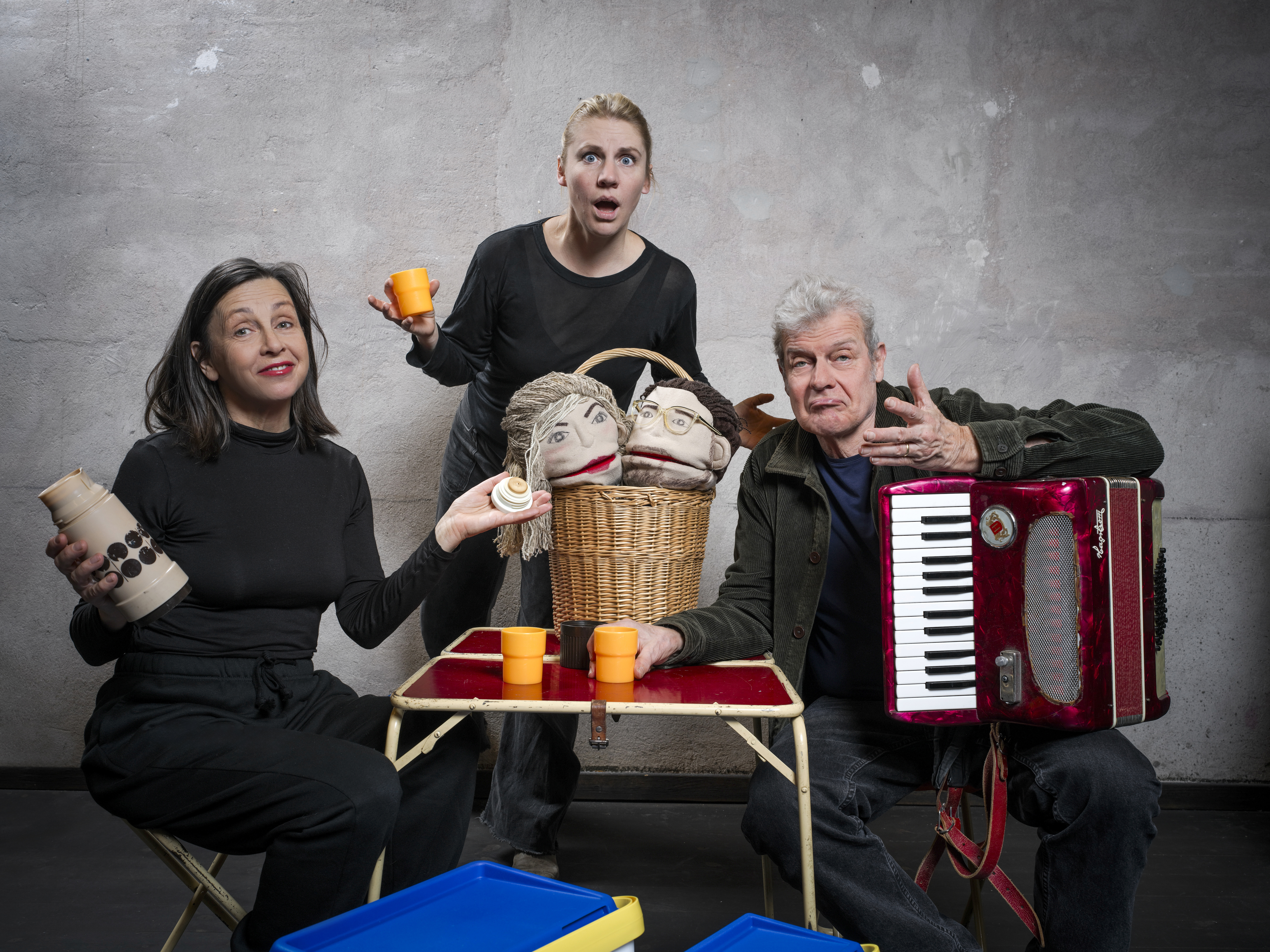 Bildtolkning: Tre personer sitter vid ett campingbord för en fikastund. EN har ett dragspel i famnen. Alla tre har ett förvånat, undrande minspel och har händerna i en frågande gest. 