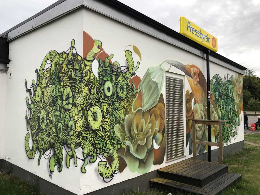 Väggmålning föreställande en fågel som håller ett munskydd i näbben, blommor och figurer.