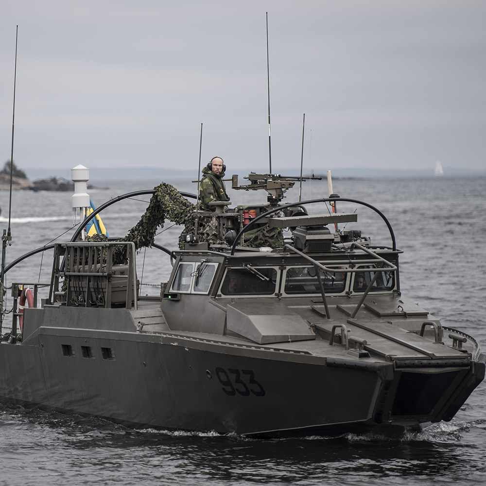 pressbild från Försvarmakten på en av marinens båtar med en soldat som tittar in i kameran och flera soldater i förarhytten